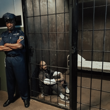 A correctional officer is standing guard outside a prison cell while an inmate sits on the floor inside.